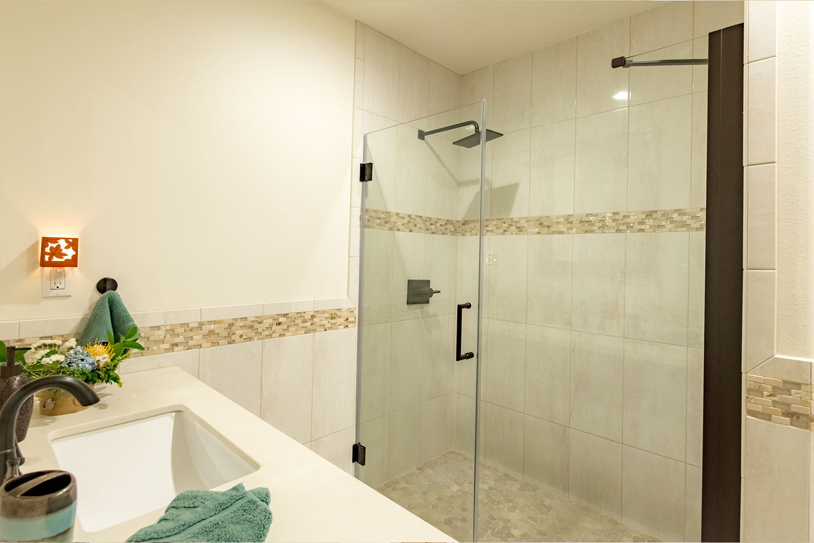 Modern bathroom featuring a white countertop, sink, black faucet, and a walk-in shower with glass doors and beige tiling. A green towel and a small potted plant are on the counter.