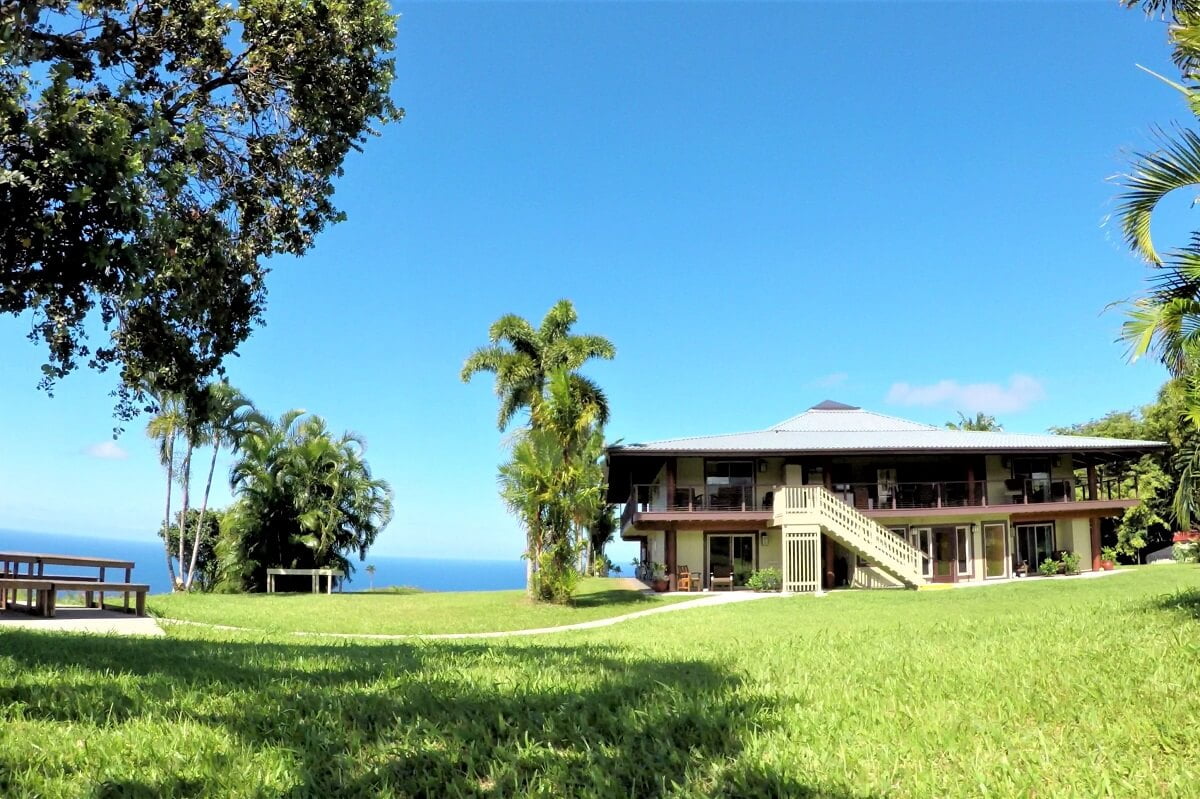 A large house with a red roof and multiple balconies overlooking a lush lawn, palm trees, and the ocean under a clear blue sky.