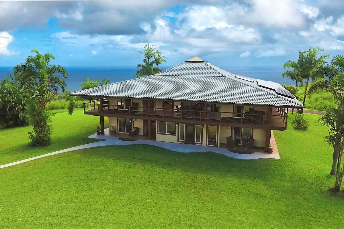 Aerial view of a large house with a gray tiled roof, surrounded by lush green grass and overlooking the ocean.