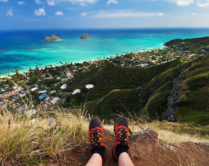 A person's legs wearing sneakers, seated on a hilltop, with a panoramic view of a coastal town and turquoise sea below.