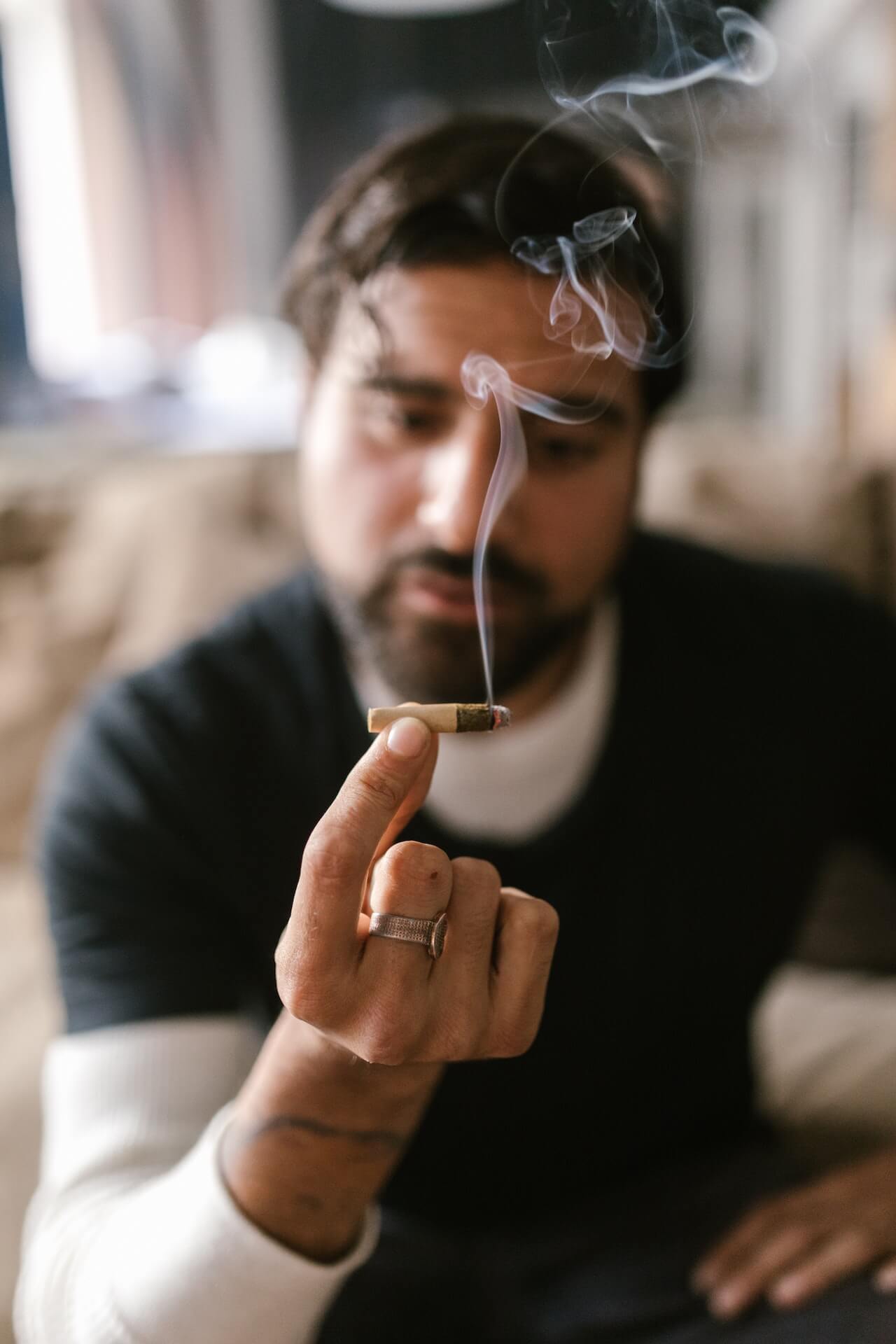 A man with a beard sits indoors, holding a smoking rolled cigarette between his fingers.