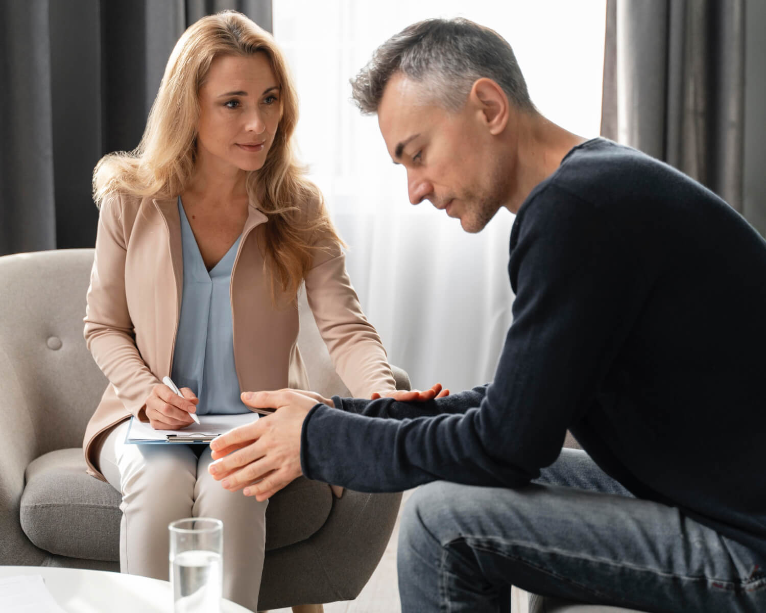 A man sits on a couch, looking down with a troubled expression, while a woman with a notepad, likely from Hawaii Rehab, sits across from him, gently touching his arm in a supportive manner.