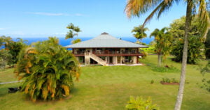 A large, two-story tropical house with a green roof, surrounded by palm trees and lush greenery, overlooks a distant ocean under a clear blue sky.