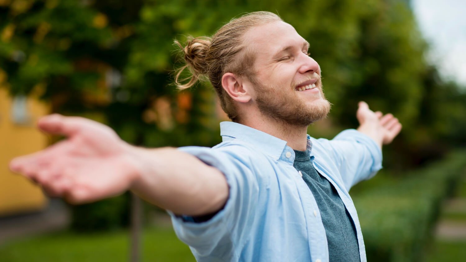 A man with a beard and a ponytail smiles with his arms outstretched, standing outdoors in a park area with trees and buildings in the background, finding joy through a non 12 step recovery program.