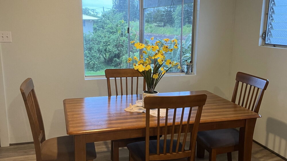 A wooden dining table with four chairs is placed near a window. A vase with yellow and white flowers is on the table. The room has a light-colored wall and wooden flooring.