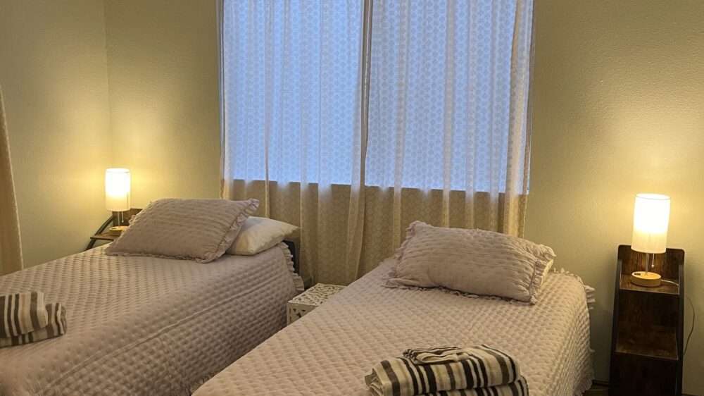 A bedroom with two single beds covered in white textured bedspreads. Each bed has a stack of striped towels on top. A small wooden nightstand with a lamp sits beside each bed.