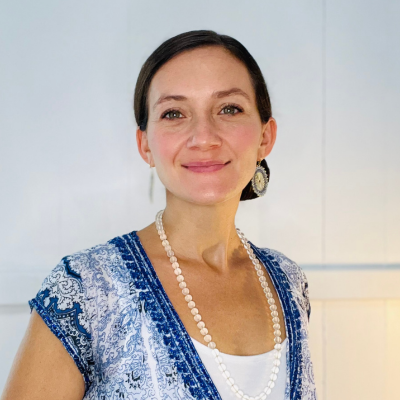 Woman with brown hair wears a blue and white patterned top, long beaded necklace, and large earring, standing against a plain white background, smiling at the camera.