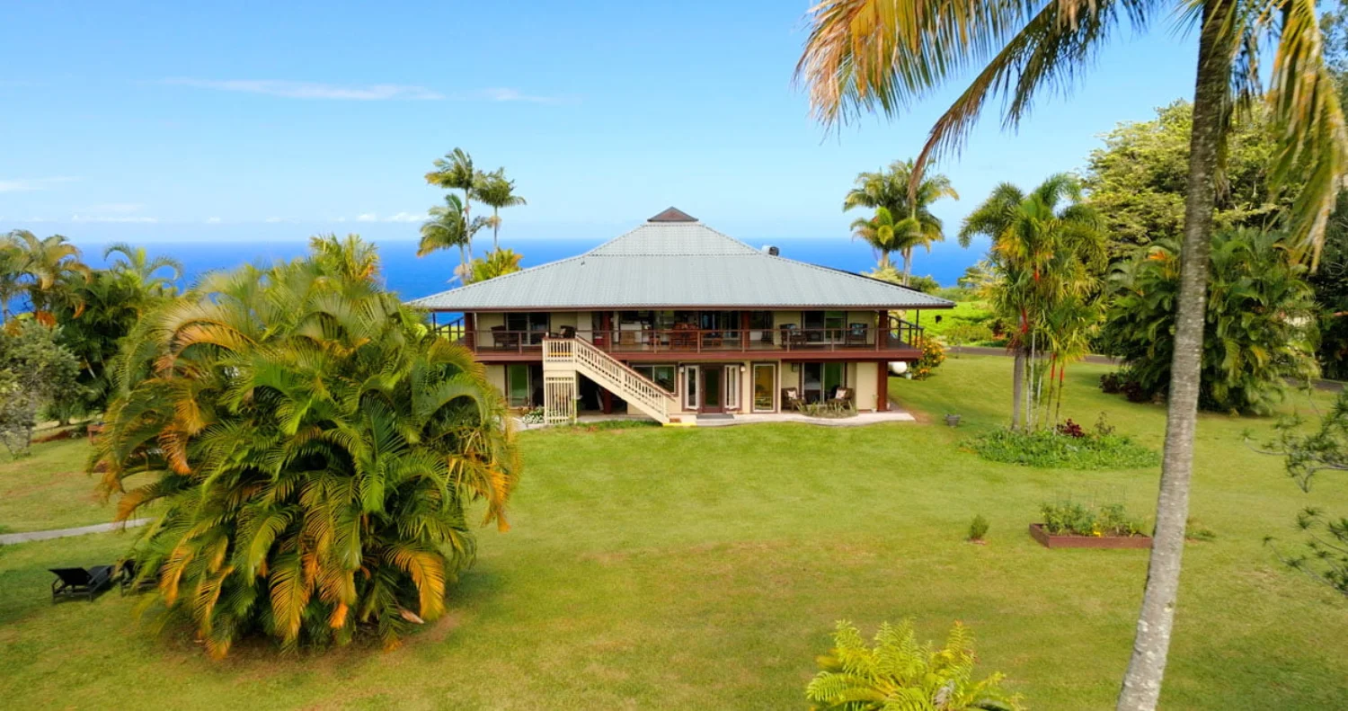A large, two-story tropical house with a green roof, surrounded by palm trees and lush greenery, overlooks a distant ocean under a clear blue sky.